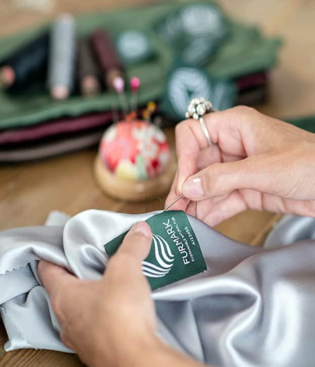 Împletire de etichetă de blană artificială pentru haine, în proces de inscripționare, cu logo-ul și detalii despre brandul FurPARK, pe o suprafață de lucru cu accesorii de croitorie și un pin colorat în fundal.